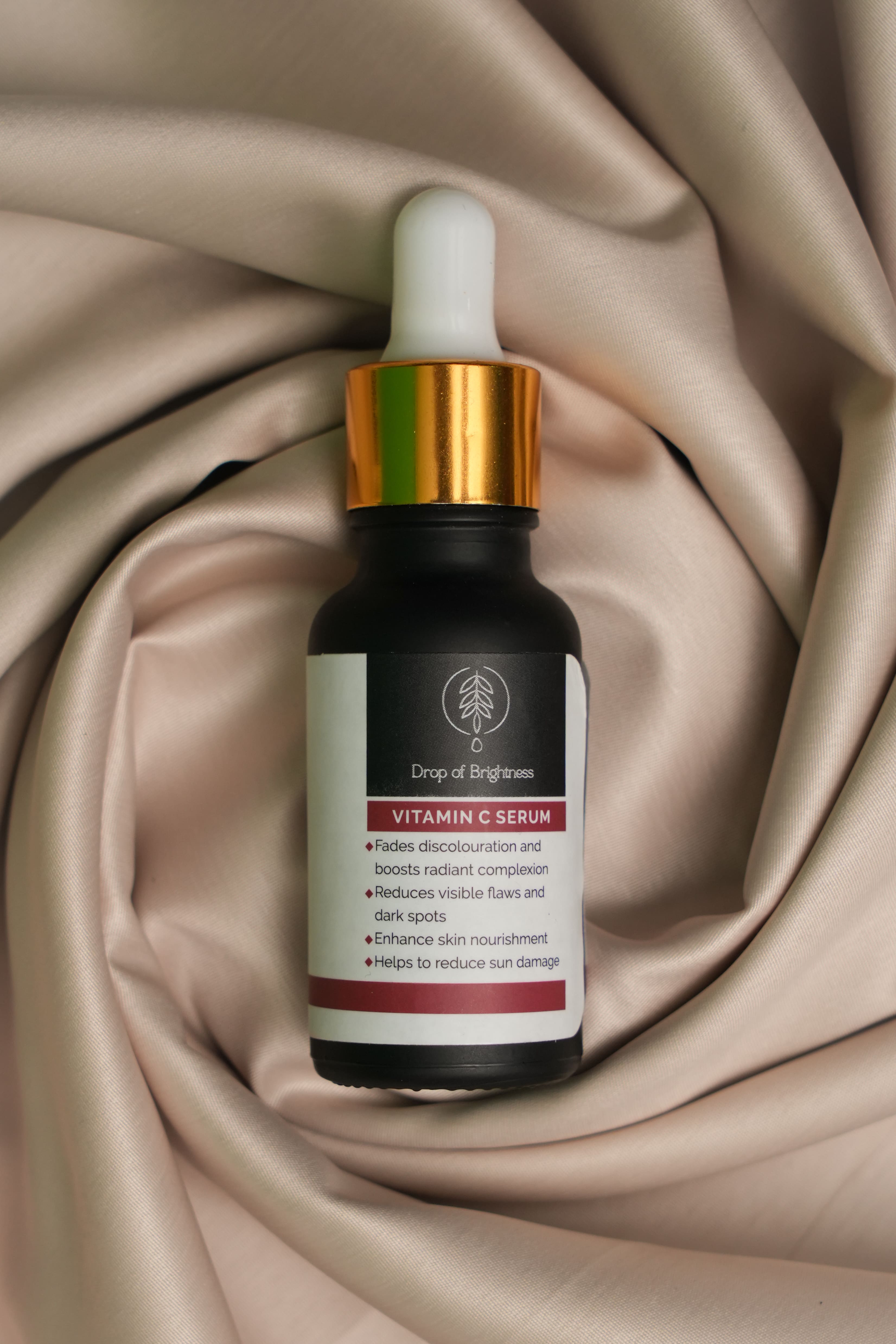 A Drop of Brightness Skin Lightening Serum by Visage Cosme, in a dropper bottle with a gold cap, sits on cream fabric. Its label claims to reduce hyperpigmentation, enhance radiance, and nourish skin for a brighter complexion.